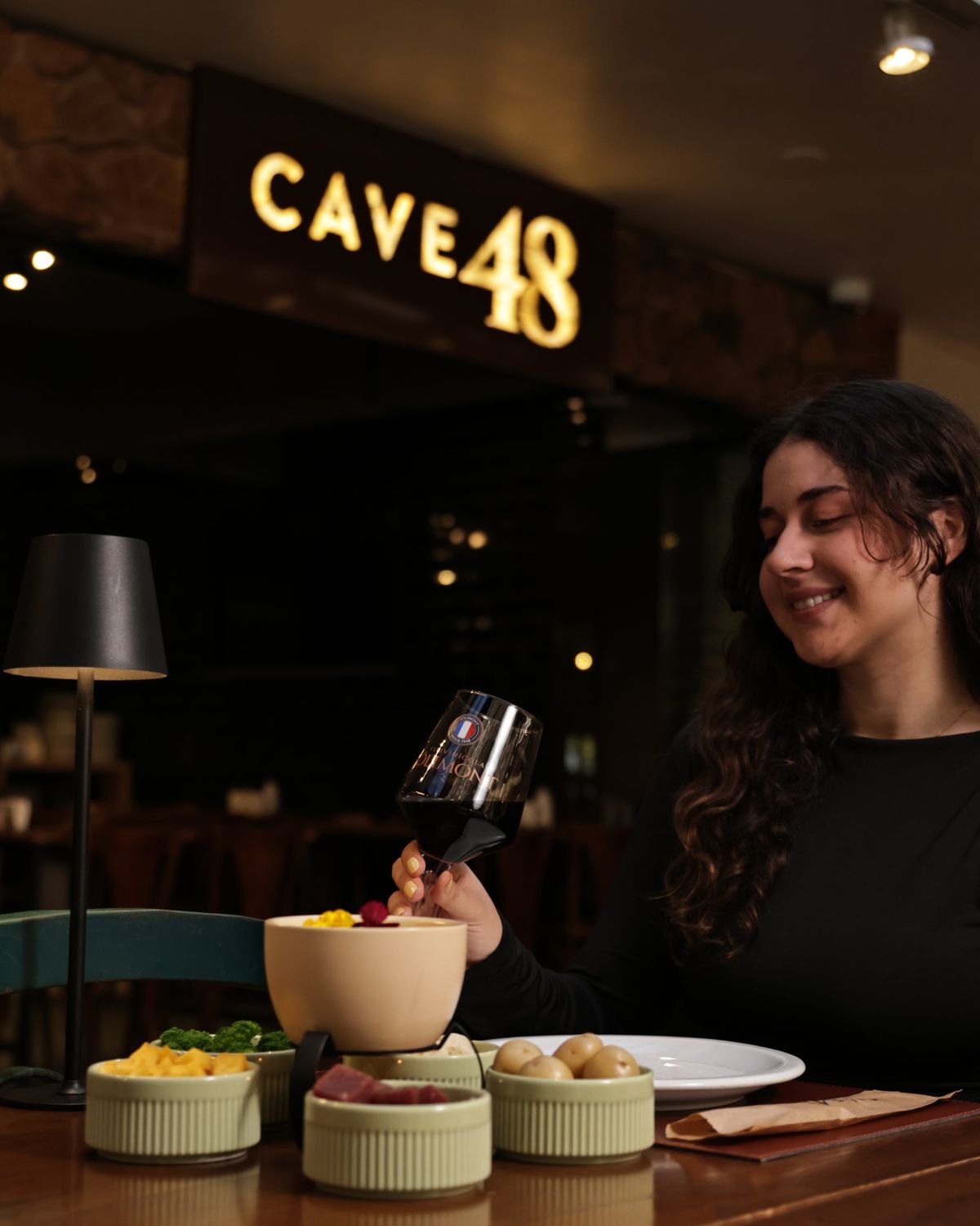Cliente brinda com uma taça de vinho Jolimont diante do fondue, sob o letreiro do Cave 48
